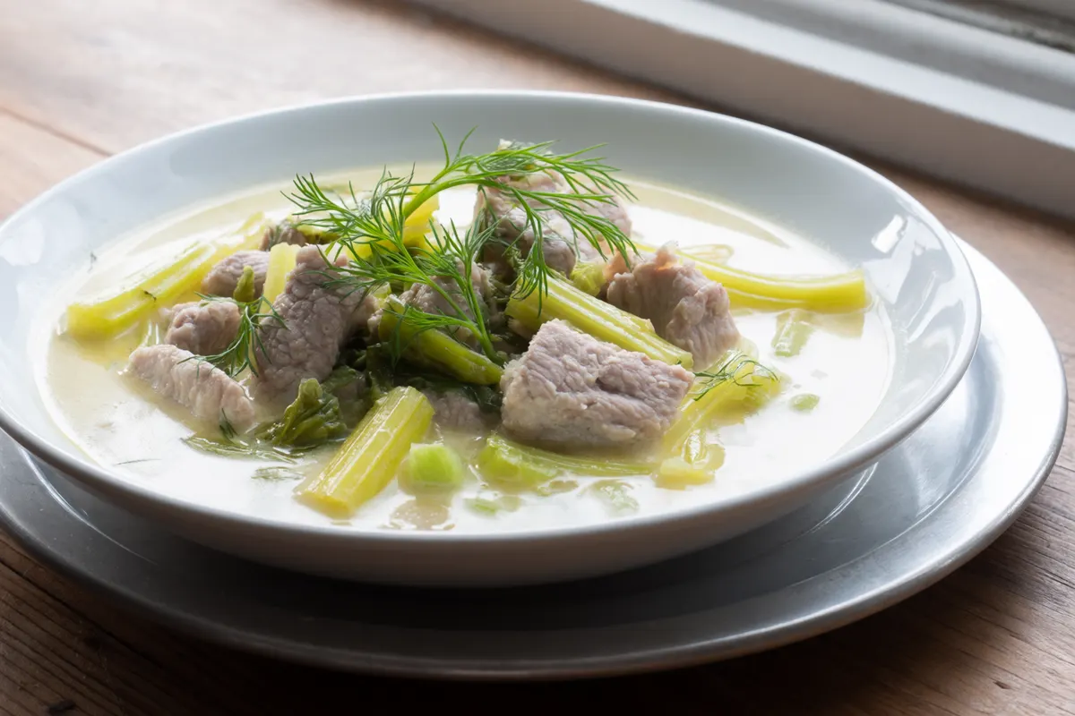 AI image of a wide white bowl of pale creamy lemon sauce with chunks of pork, celery stalk pieces, celery leaves and a sprig of dill.